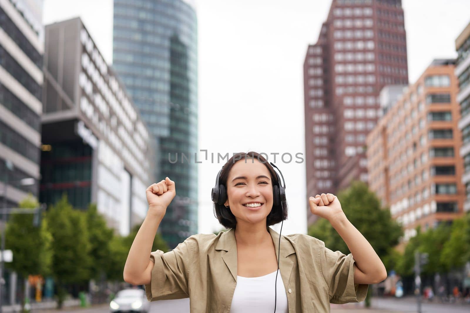 Portrait of smiling asian woman dancing, triumphing, feeling happy while listening music in city, posing on street near skyscrappers.