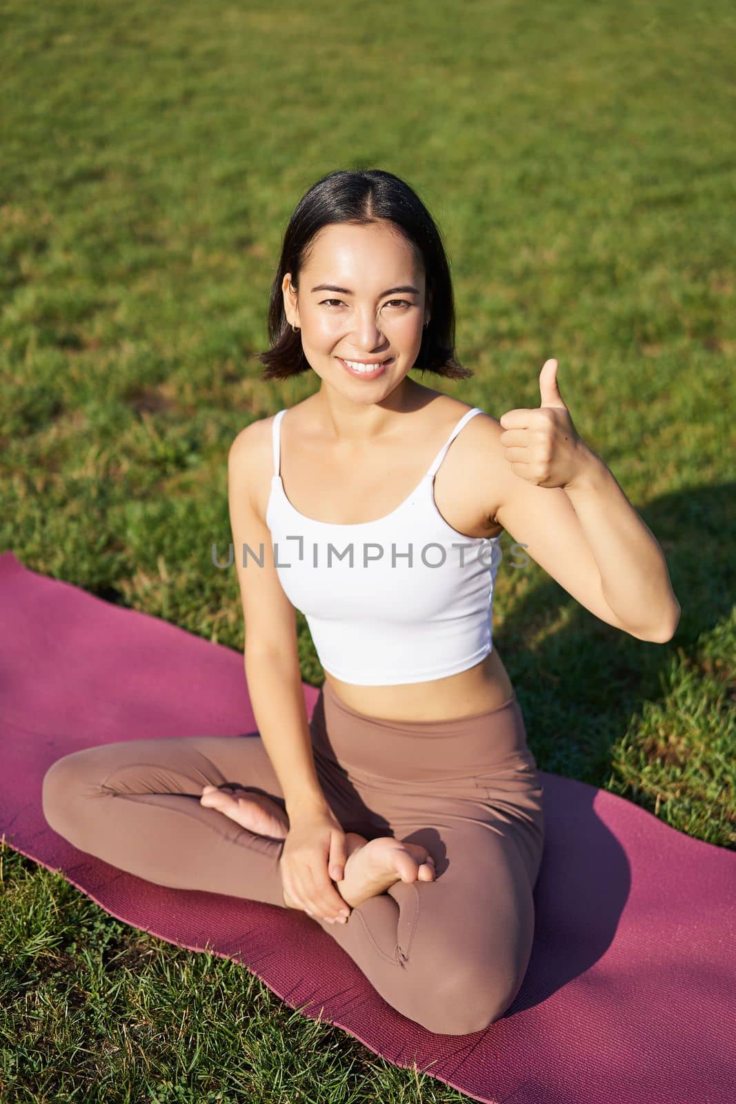 Vertical shot of asian woman sitting on rubber mat after good yoga training in park, showing thumb up in approval. Meditating on fresh air.