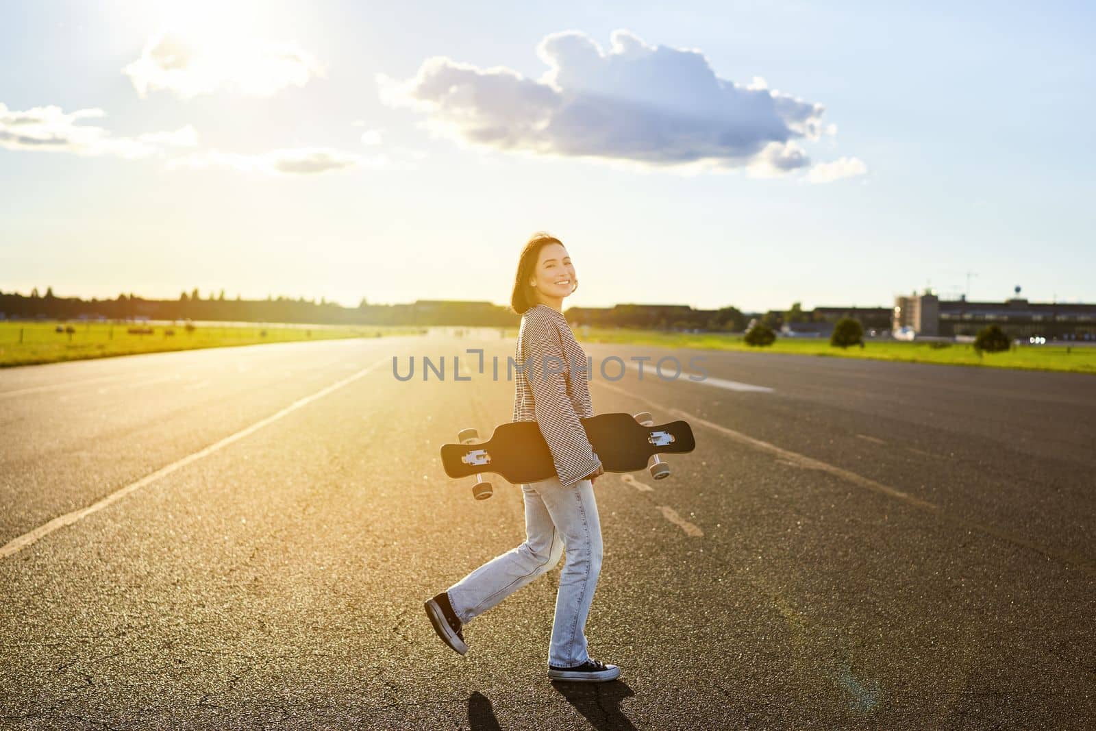 Asian girl with skateboard standing on road during sunset. Skater posing with her long board, cruiser deck during training by Benzoix