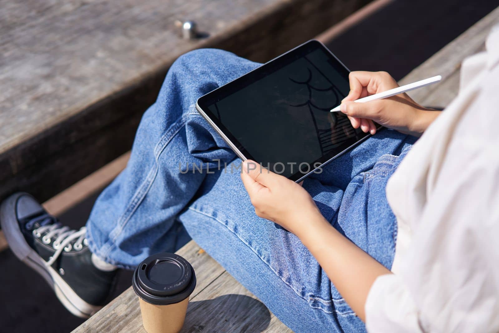 Cropped shot of female hands drawing on digital graphic tablet with pen, sitting with cup of coffee outdoors on bench by Benzoix