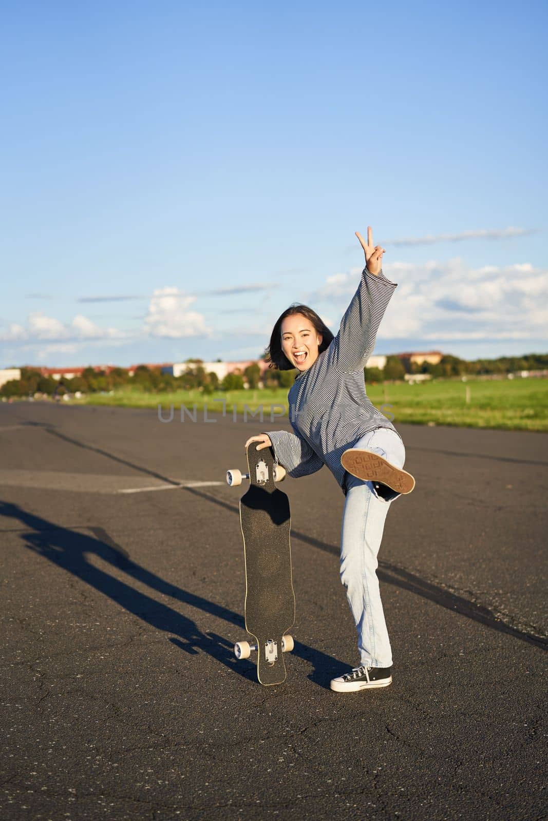 Vertical shot of happy asian skater girl, jumping, standing with skateboard and smiling. Woman skating on longboard and having fun.