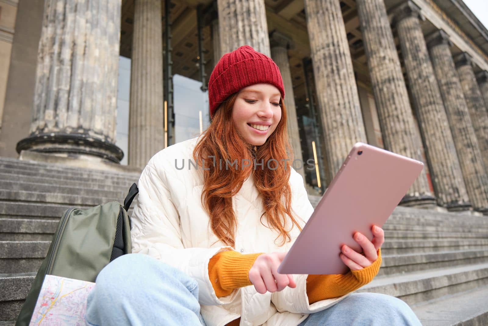 Outdoor shot of young stylish redhead girl sits on staircase and connects to public wifi, uses digital tablet, reads news on gadget by Benzoix