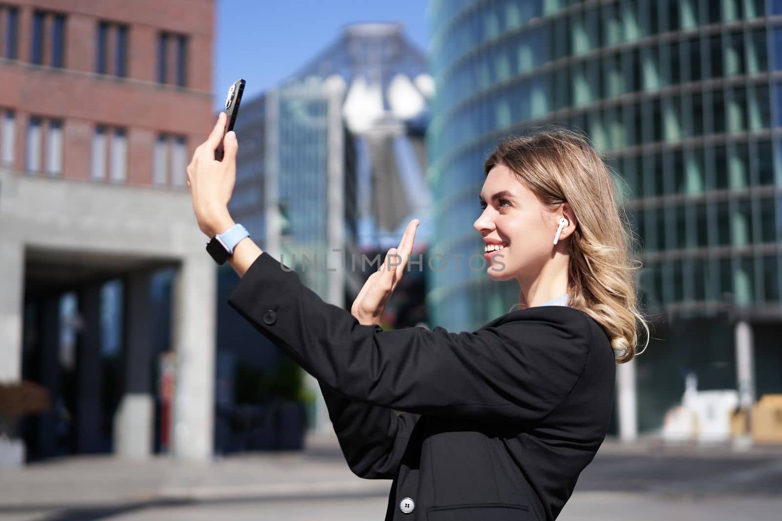 Portrait of businesswoman wave her hand at mobile phone camera, waves hand during video chat, stands in suit in city center outdoors.