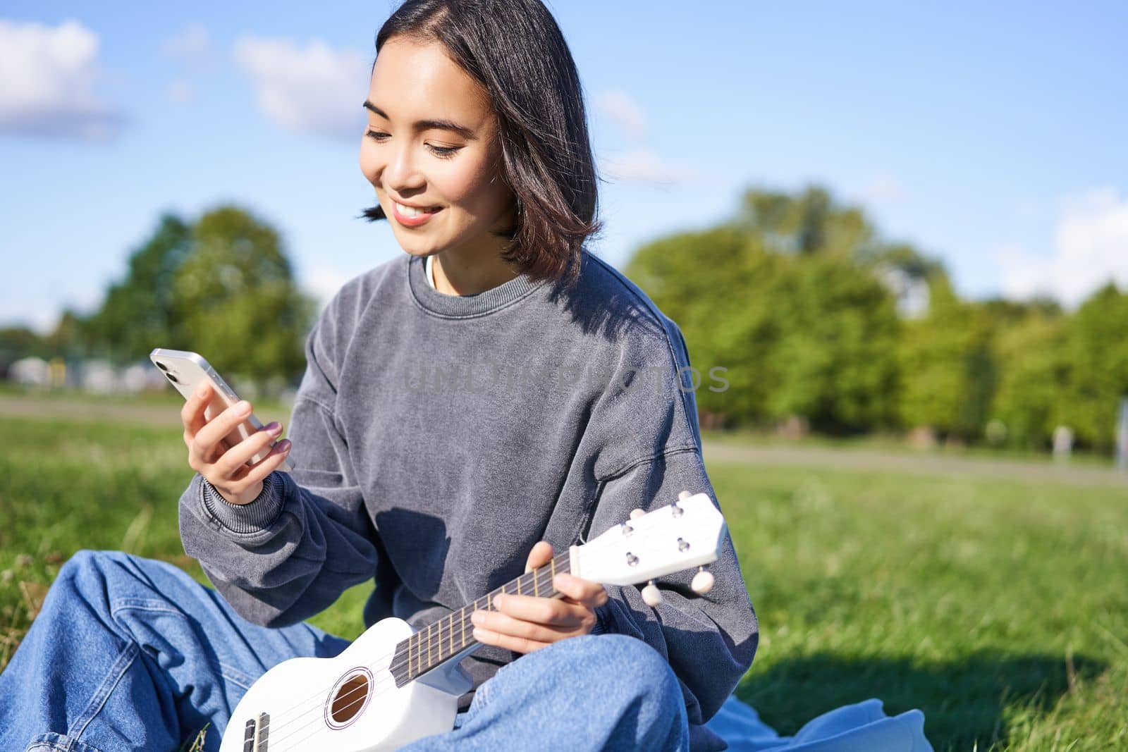 Technology and music. Smiling asian hipster girl sitting in park with smartphone and holding ukulele, playing instrument.