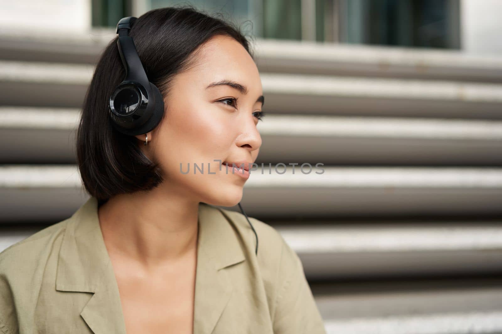 Young asian woman listening to music in headphones, sitting outside on street, smiling.