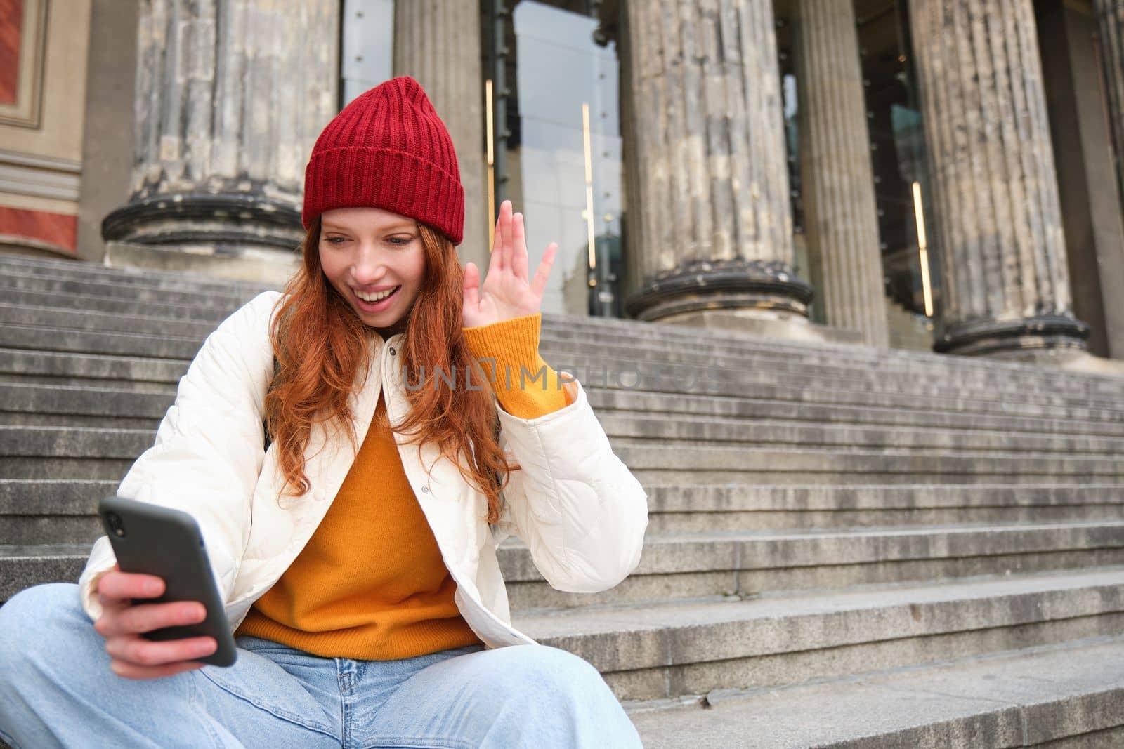 Young redhead woman sits on stairs outdoors and waves hand at smartphone camera, video chats with friends, connects to public wifi by Benzoix