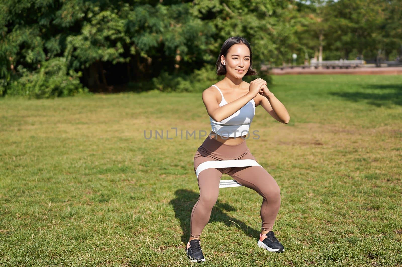 Woman using stretching band, resistance fitness rope for workout in park, doing squats, training on fresh air by Benzoix