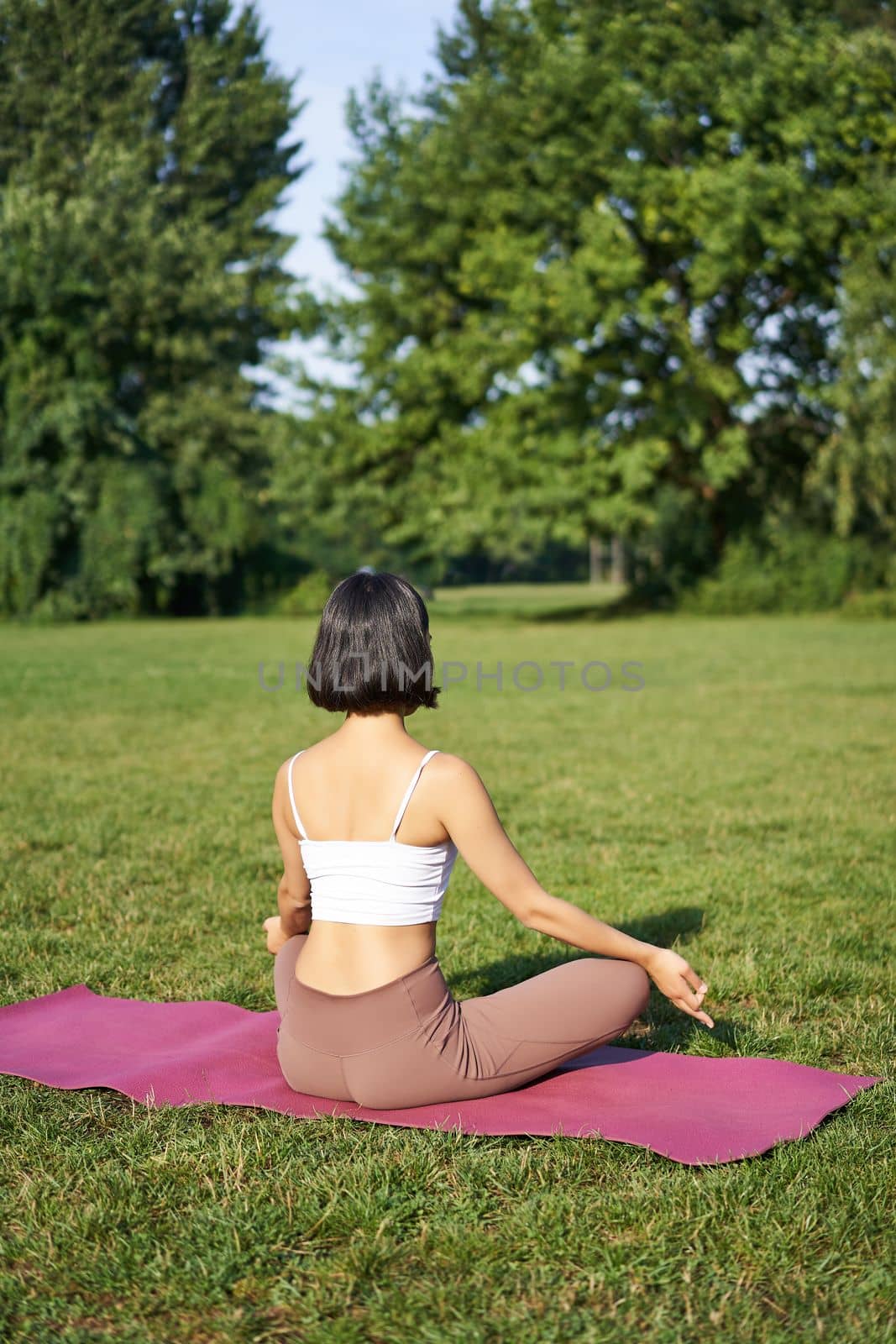 Rear view of woman silhouette doing yoga, sitting on fitness mat and meditating on green lawn.