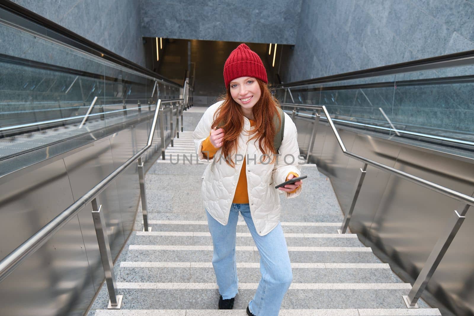 Portrait redhead girl tourist, goes up stairs with smartphone, follows route on mobile phone app, holds backpack and smiles.