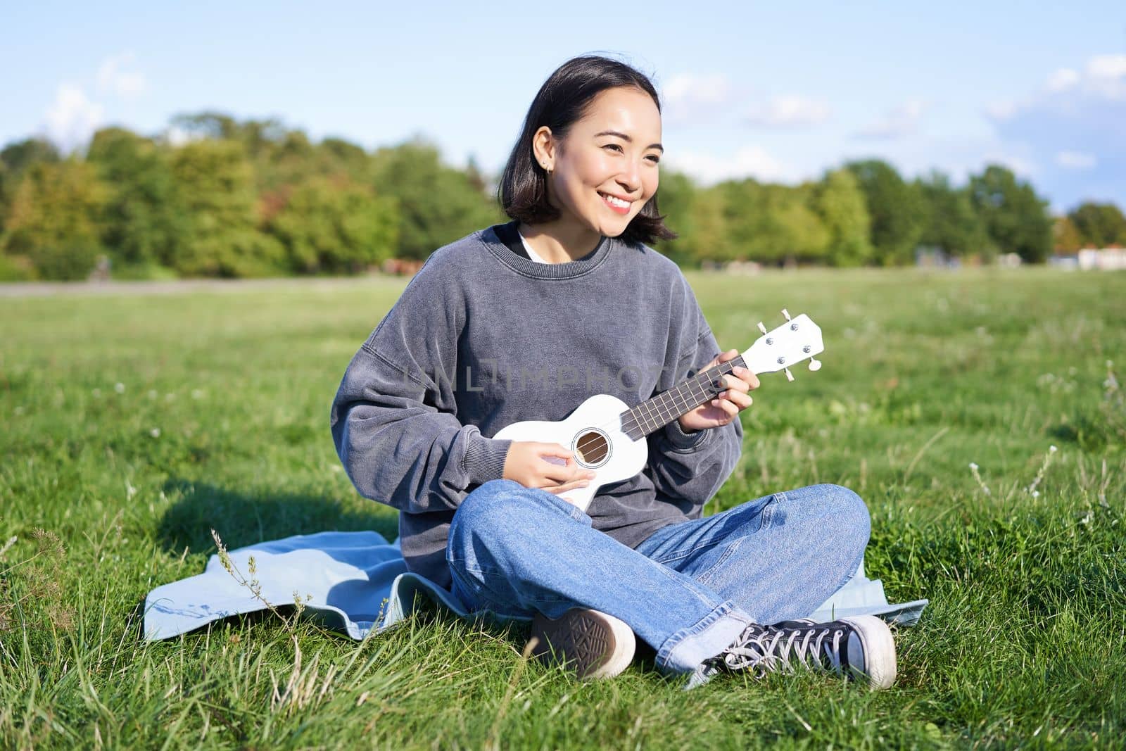 Beautiful asian girl sitting in park, playing ukulele and singing, relaxing outdoors on sunny spring day.