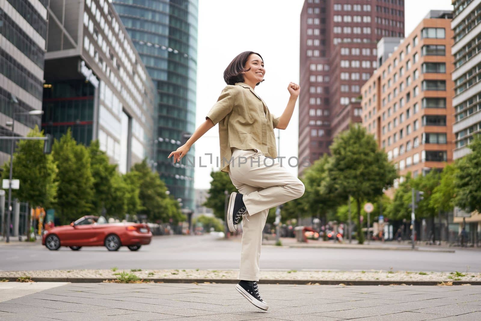 Portrait of asian happy girl jumping and dancing in city centre, posing on streets, express joy and excitement by Benzoix