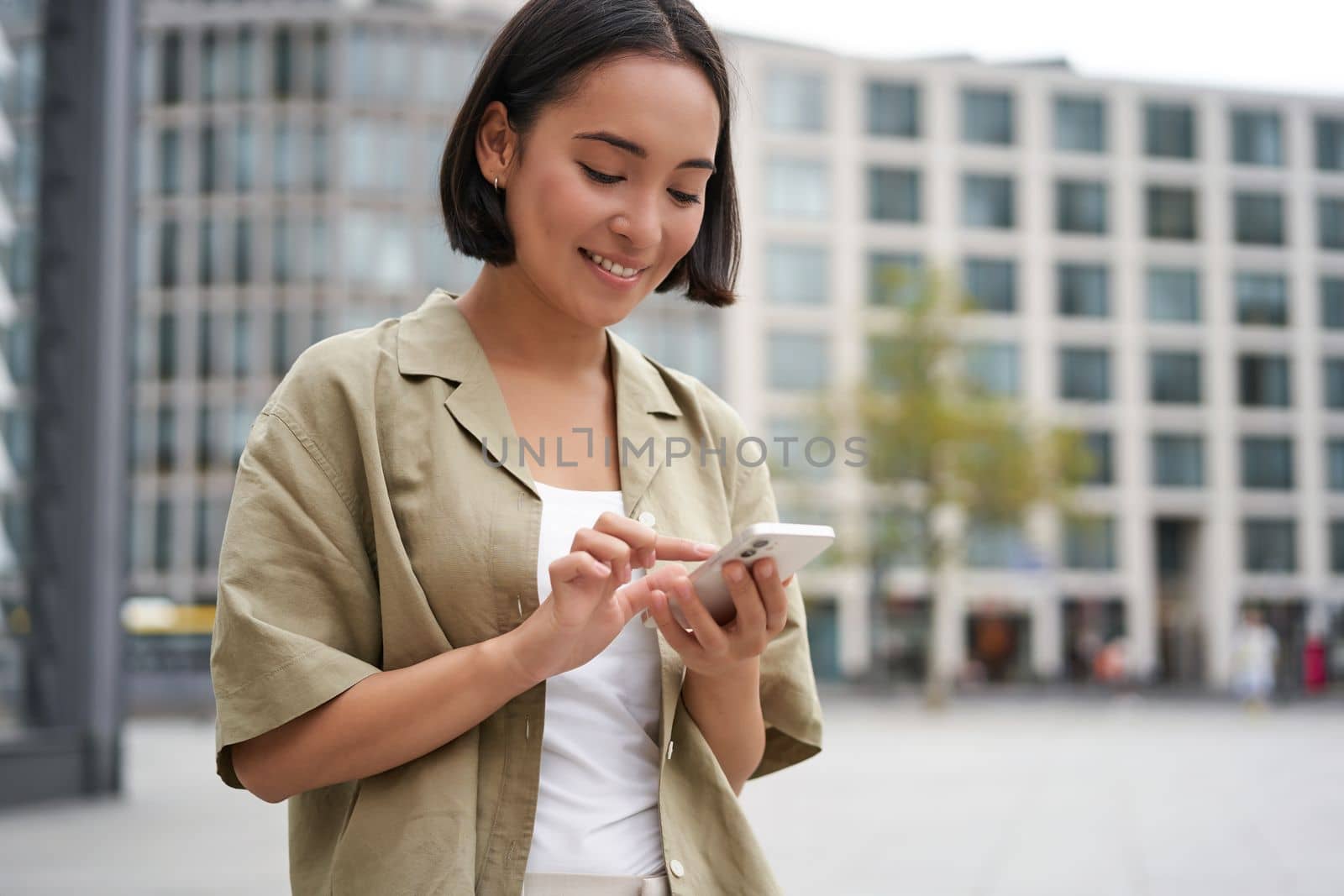 Mobile technology. Smiling asian woman using smartphone app, looking at her telephone on street, checking map, calling or texting someone by Benzoix