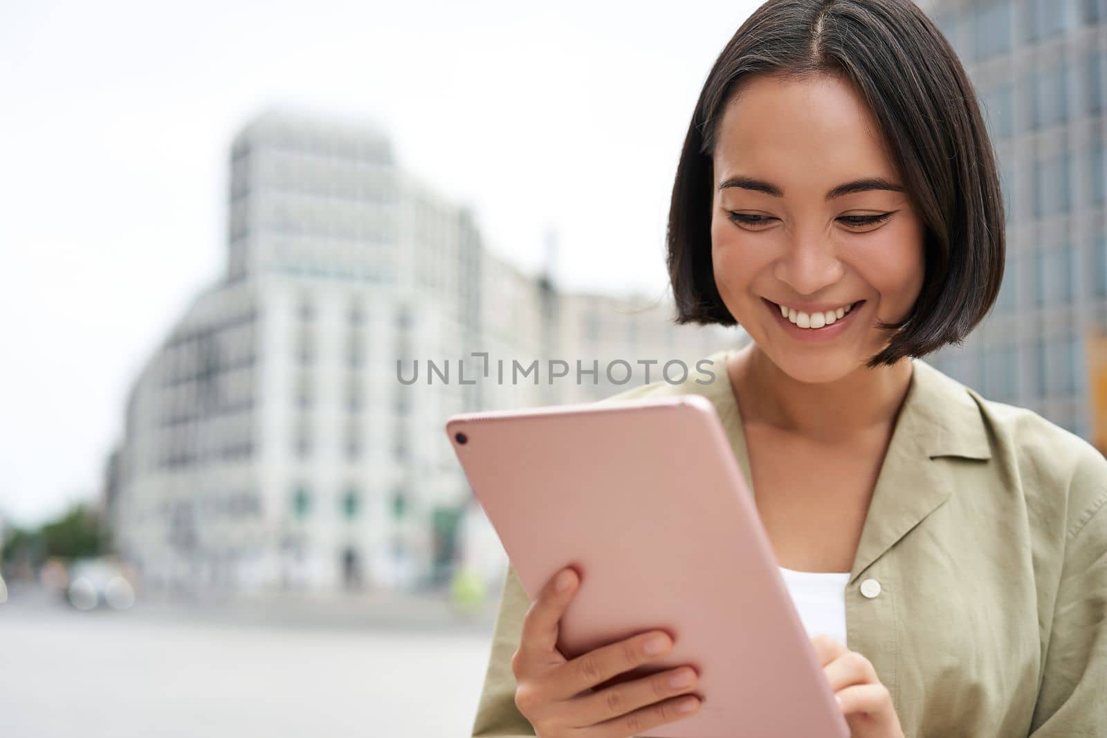 Portrait of asian woman reading, using tablet while standing on street, smiling while looking at screen by Benzoix