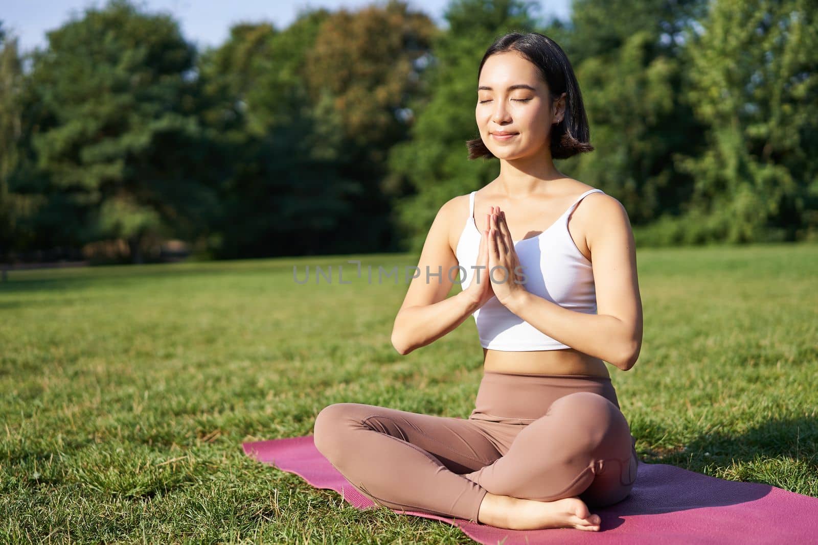 Woman meditating on lawn in park, sitting on sports mat, relaxing, breathing fresh air.