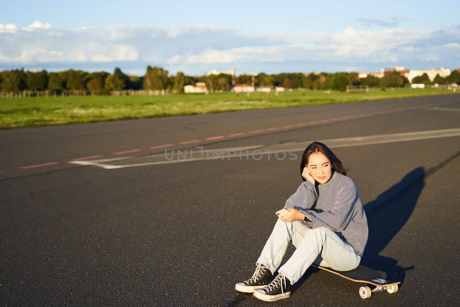 Skater girl sits on her skateboard on road, using smartphone, chatting on mobile app.
