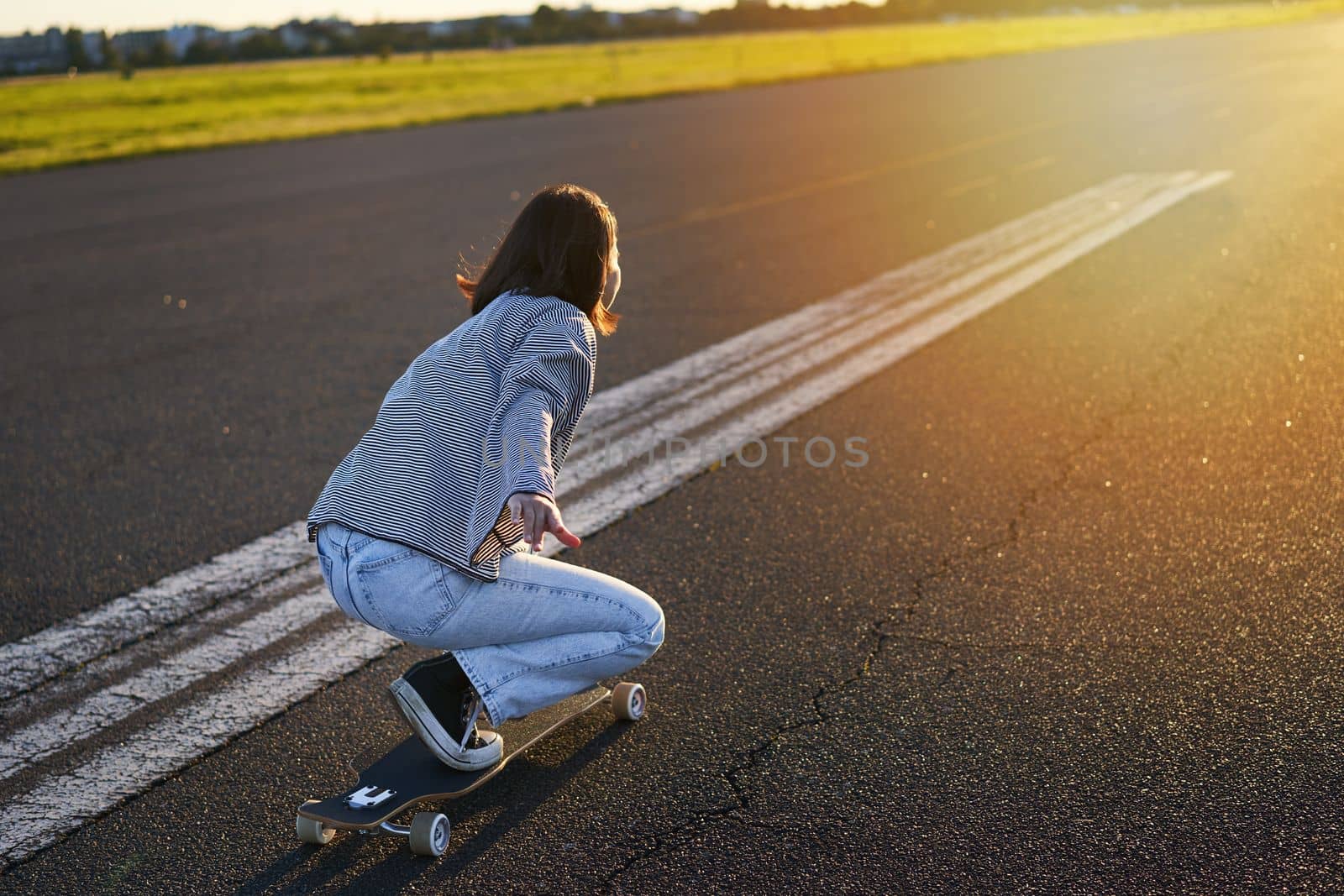 Side view of beautiful asian girl on skateboard, riding her cruiser towards the sun on an empty road. Happy young skater enjoying sunny day on her skate.