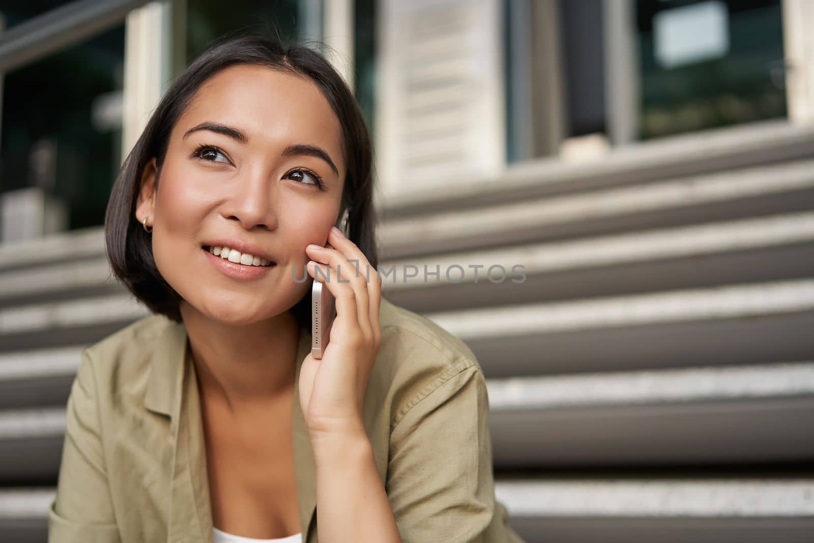 Cellular technology. Smiling asian girl talks on mobile phone, makes a call. Young woman with telephone sits on stairs by Benzoix