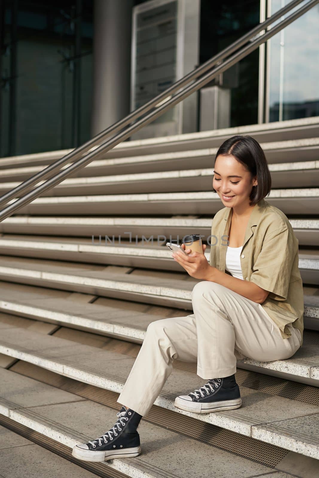 Vertical shot of young smiling woman sits on stairs in city centre wth mobile phone. Girl with telephone, browsing on smartphone, texting message by Benzoix