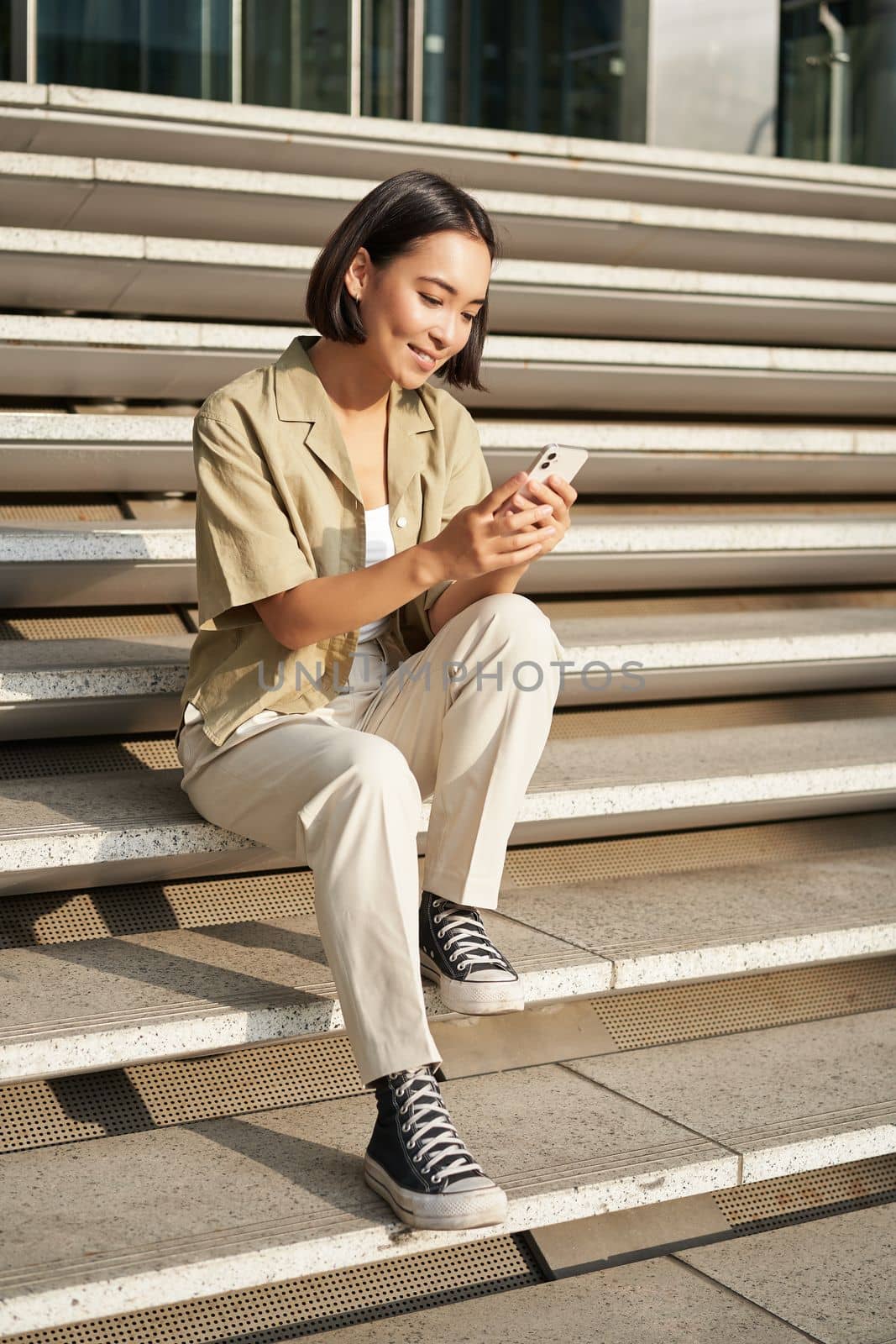 Beautiful asian girl sitting on stairs outside building, using mobile phone, looking at smartphone app and smiling.
