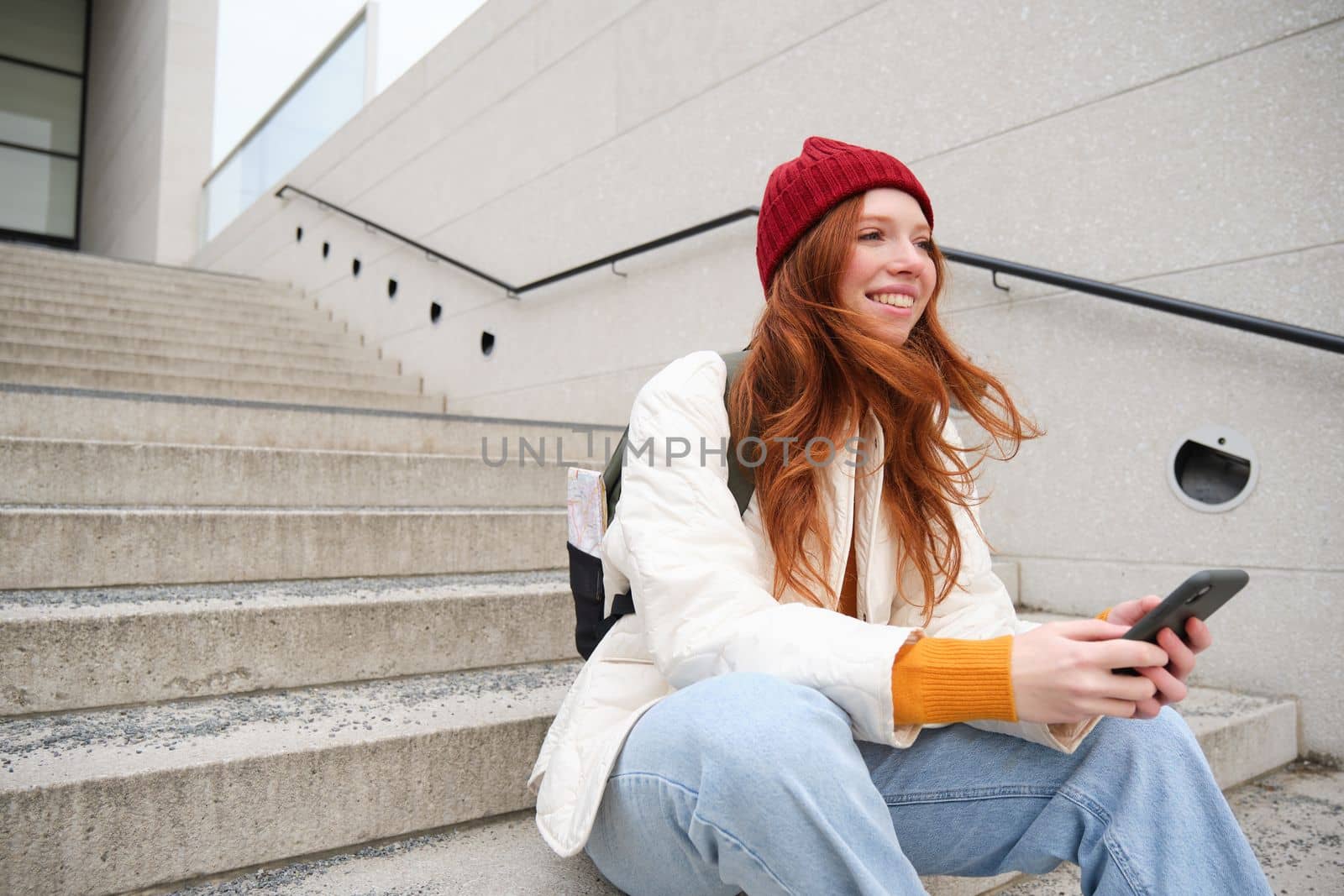 Mobile phones and people. Young stylish redhead girl sits on stairs with telephone, uses smartphone app, reads smth online by Benzoix