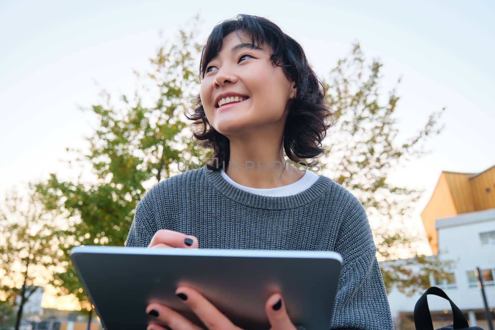 Close up portrait of young korean girl sits outdoors in park, holds her digital tablet and graphic pen, draws scatches, gets inspiration from nature for art, smiles happily by Benzoix