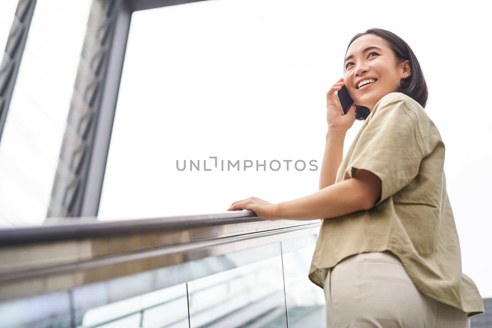 Happy asian woman on escalator, talking on mobile phone, meeting someone in city by Benzoix