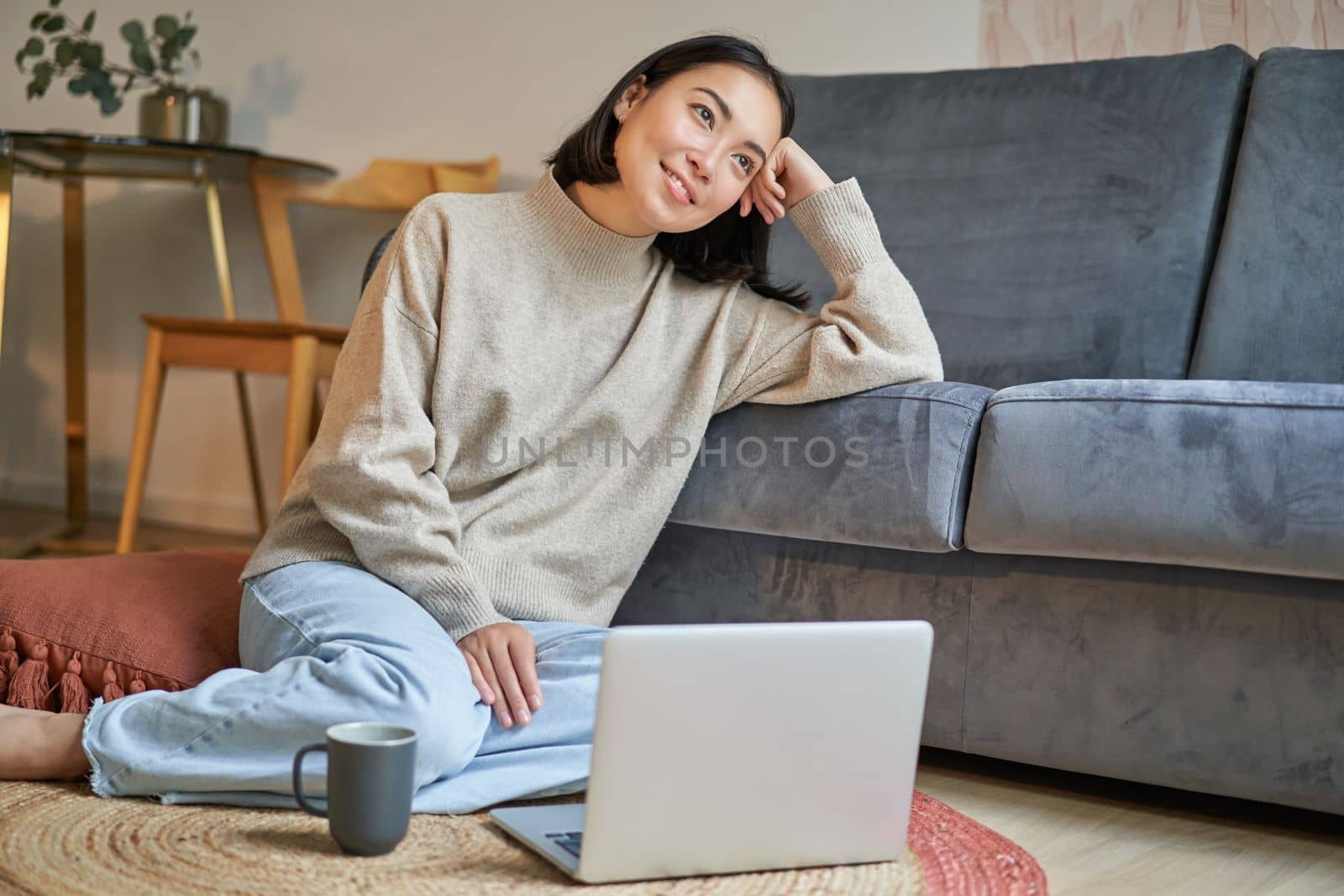 Portrait of dreamy woman sitting with laptop on floor, watching on computer and drinking coffee, enjoying cozy days at home.