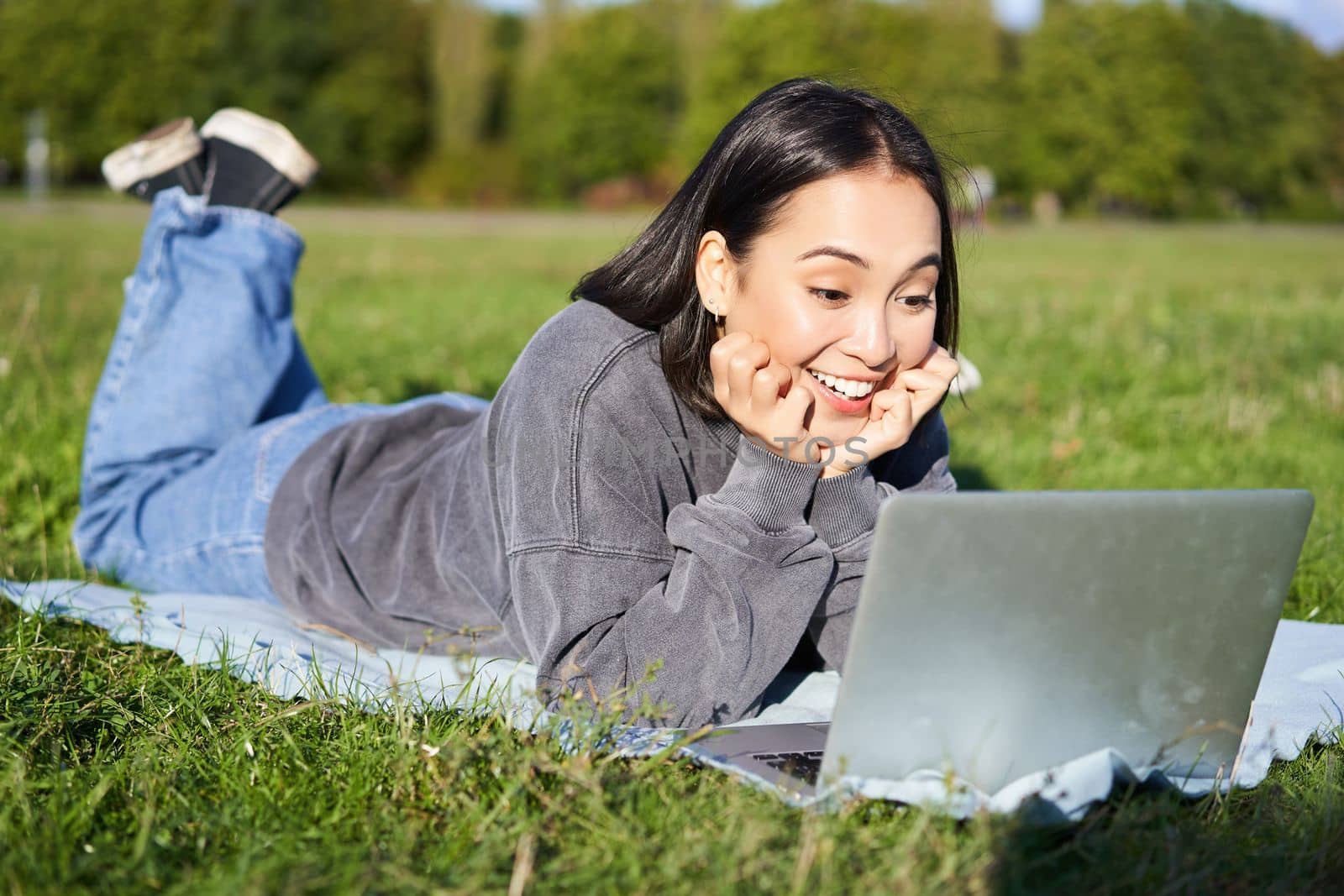Portrait of beautiful girl lying in park and looking with surprised face at her laptop, watching videos, chatting with friends while relaxing outdoors by Benzoix