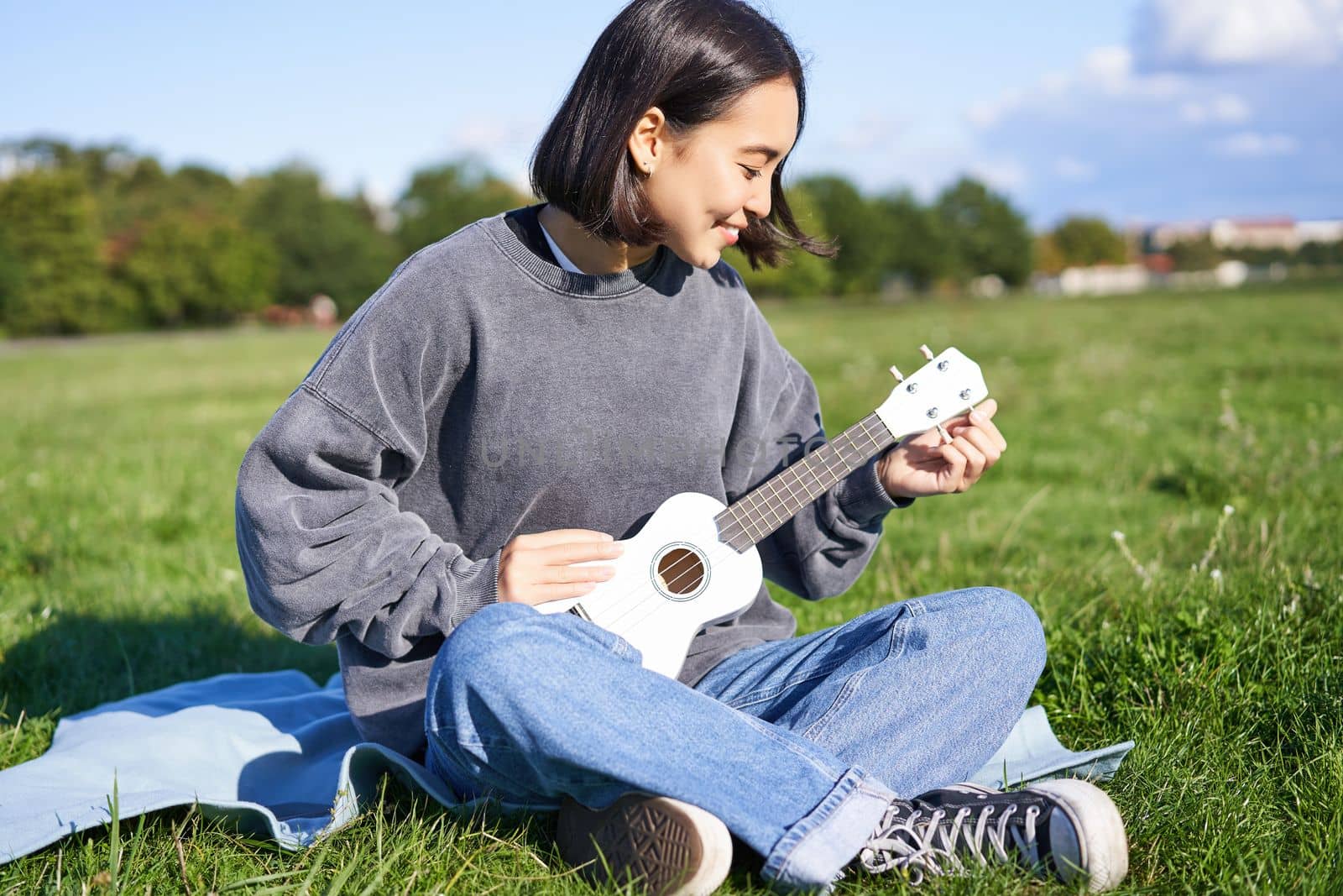 Asian smiling girl tuning her ukulele guitar, singing and playing in park, sitting on blanket on sunny day.