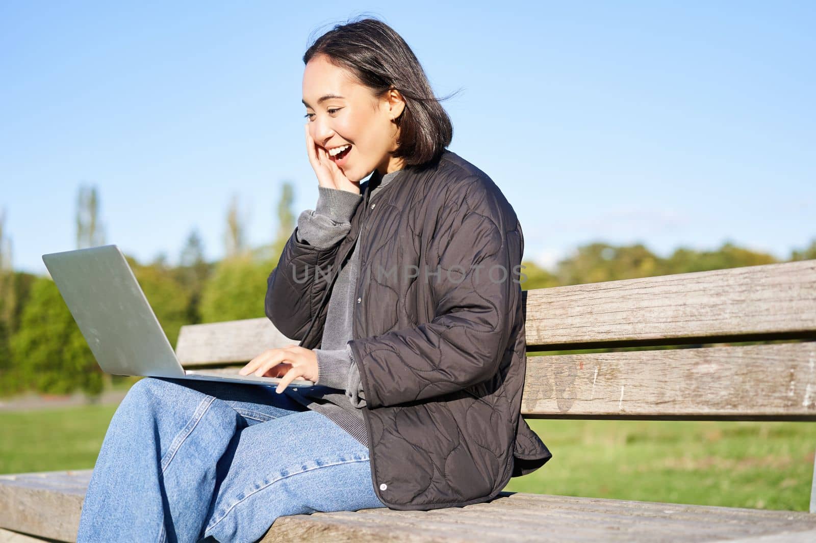 Portrait of smiling asian girl sits on bench in park, talks to friend online via laptop, video chats, using computer to study on remote.