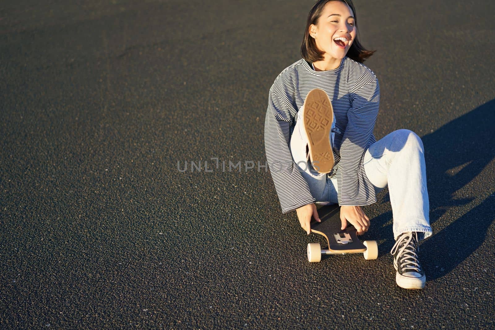 Portrait of beautiful asian girl skating, sitting on her skateboard and smiling. Cute teenager with longboard, skating on road.