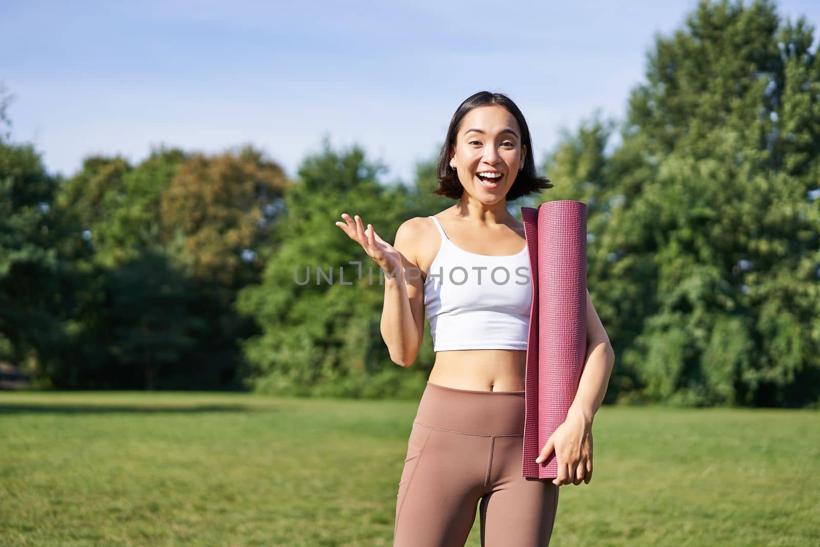 Excited asian girl in sportswear, holds rubber mat for yoga, looks surprised and happy, stands in park, workout on fresh air outdoors.