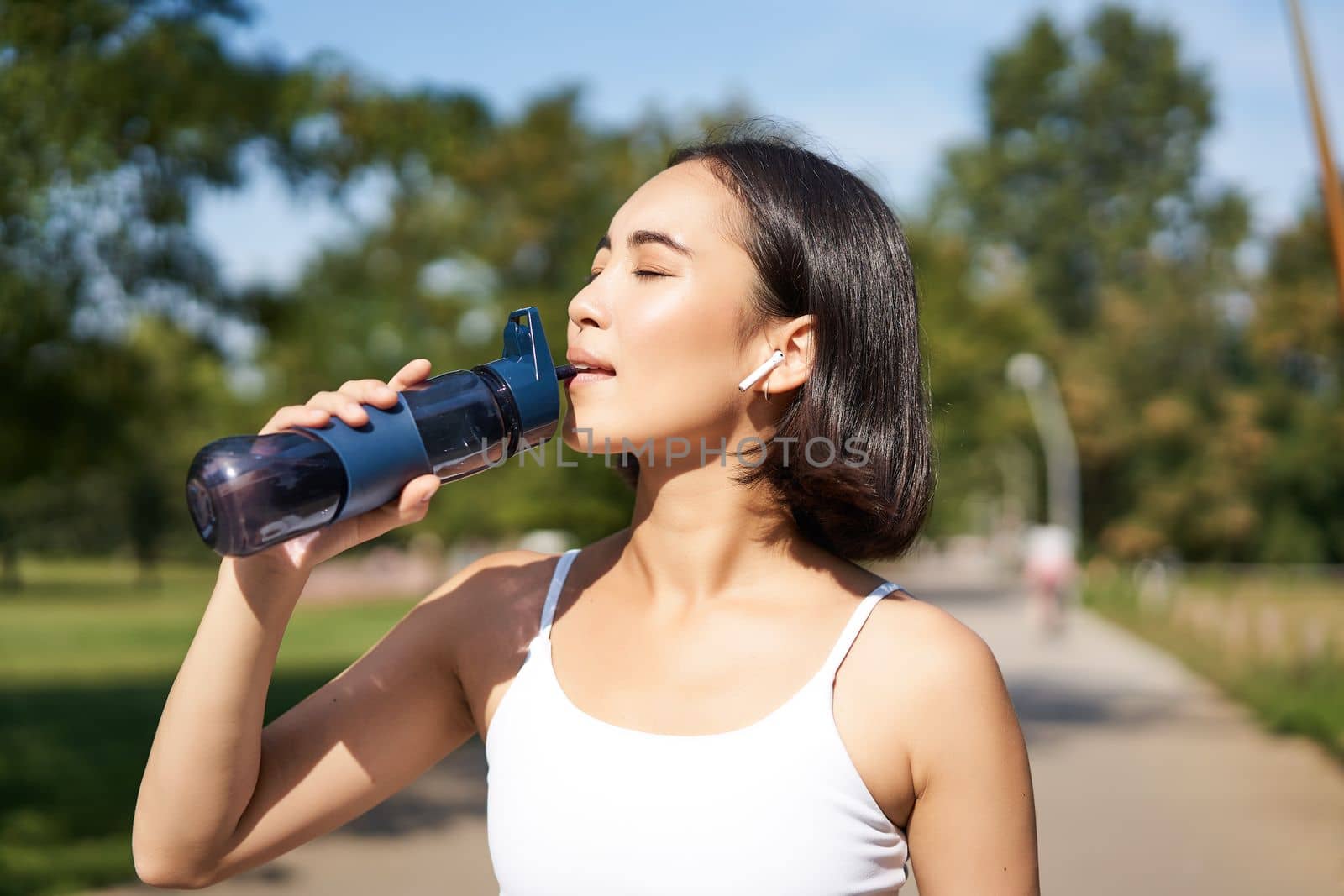 Happy asian sportswoman, runner drinks water from bottle while running, workout on fresh air in park.