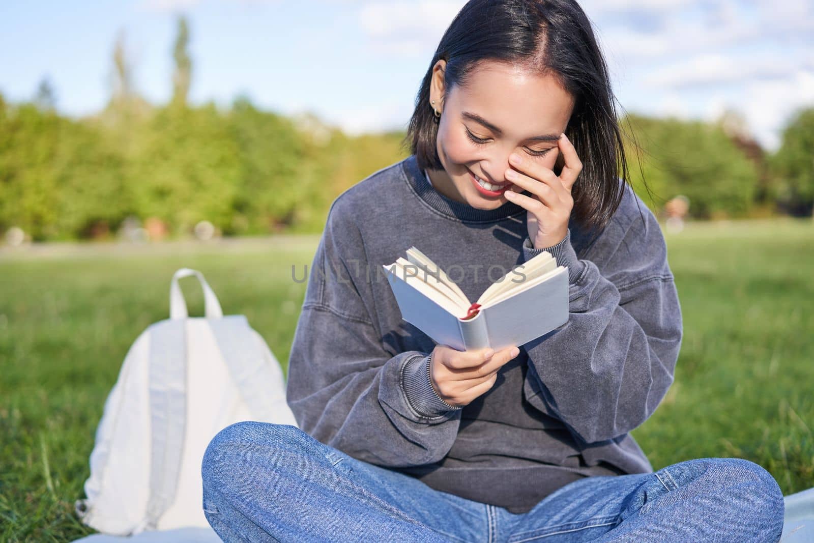 Beautiful woman sitting in park with book. Smiling asian girl reading and laughing, relaxing outdoors.