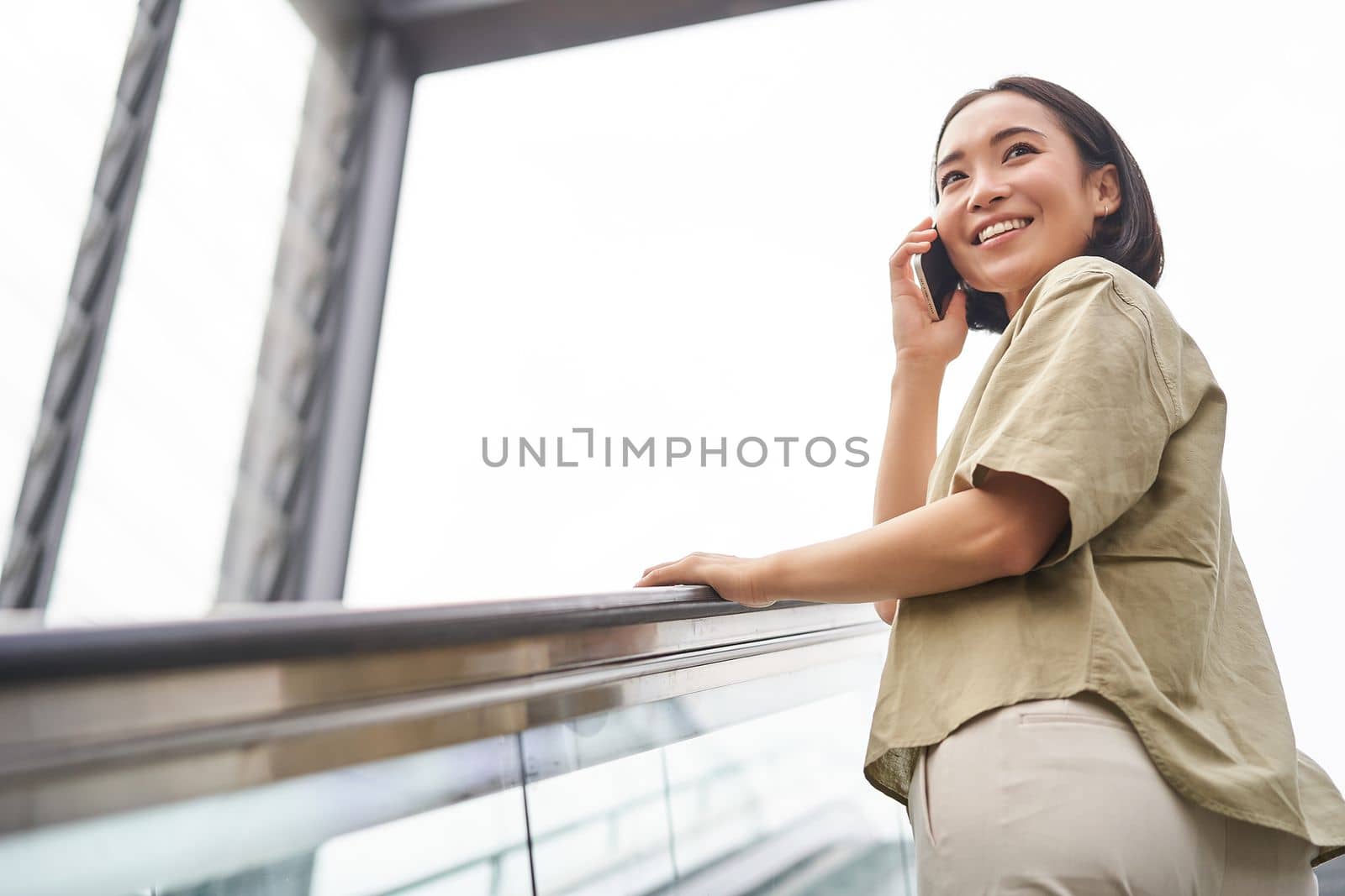 Happy asian woman on escalator, talking on mobile phone, meeting someone in city. Copy space