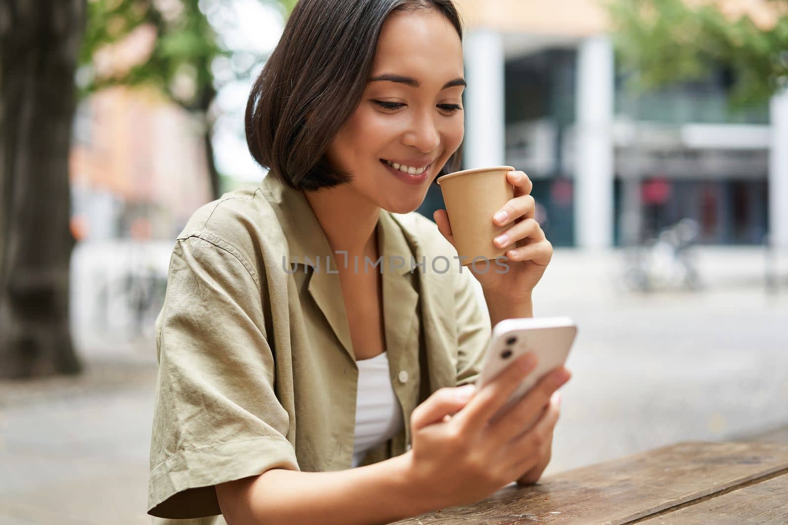 Close up portrait of asian girl sitting in cafe outside, drinking coffee and using smartphone, reading mobile phone.