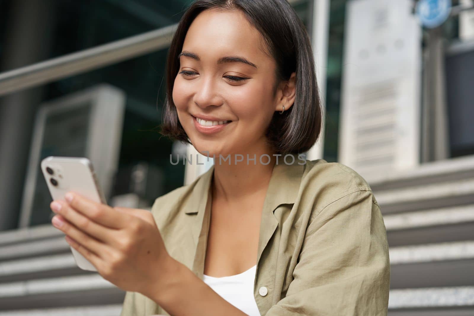 Portrait of asian girl takes selfie on mobile phone. Korean woman smiling, video chat on smartphone app while sits outside on stairs by Benzoix