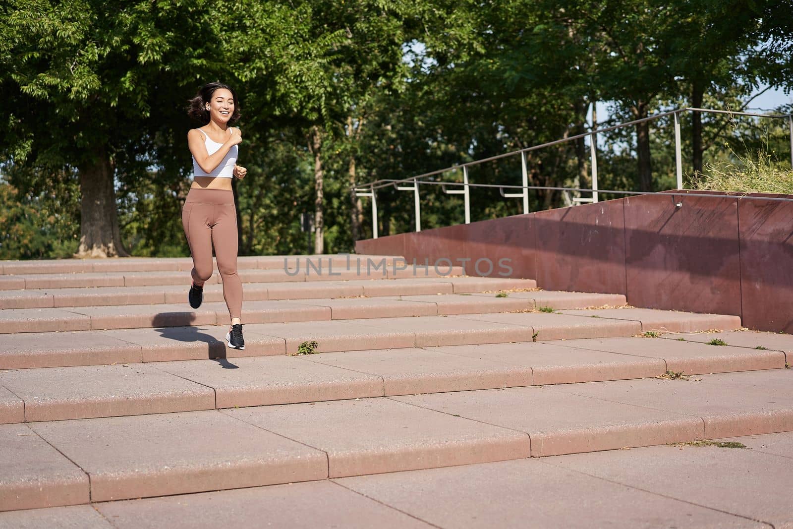 Healthy fitness girl running outdoors on street, wearing uniform, jogging on fresh air and listening music in wireless headphones.