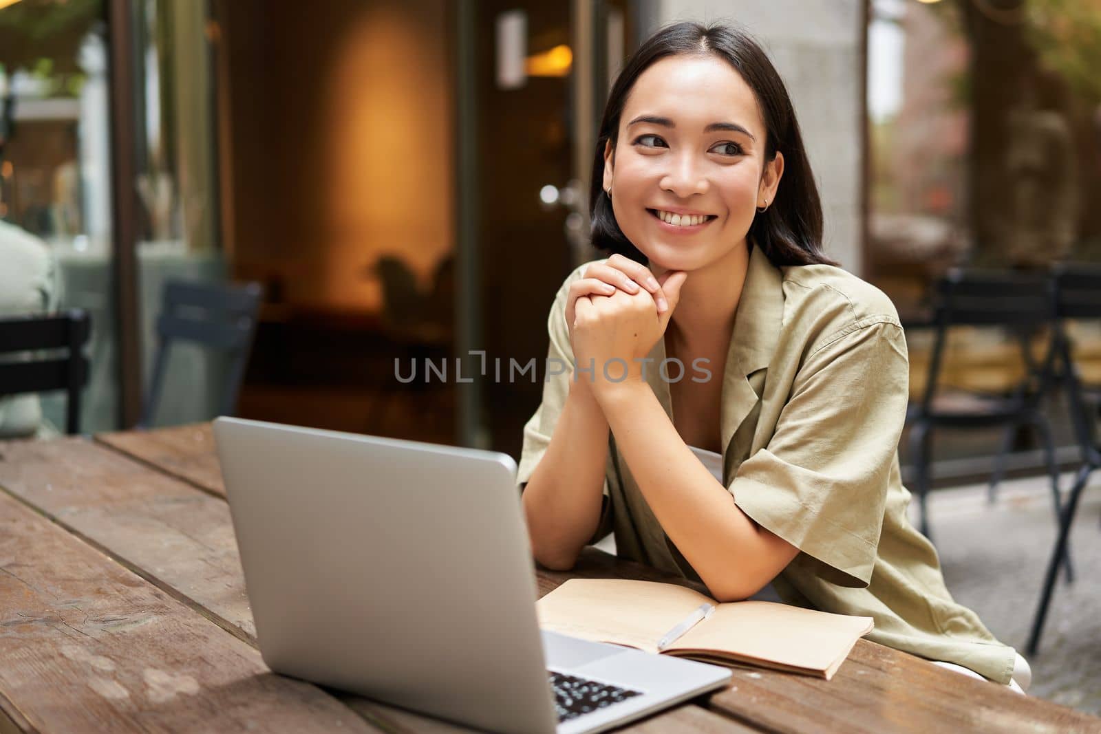 Portrait of asian girl works outdoors in cafe, sits with laptop, studies, smiles happy.