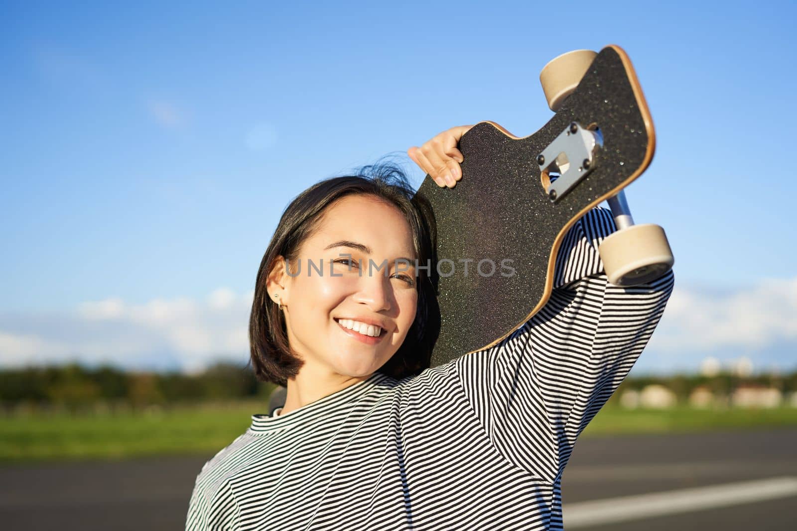 Happy and free asian girl holding cruiser board on shoulders and walking towards camera on empty road, skating on longboard and enjoying sunny weather by Benzoix