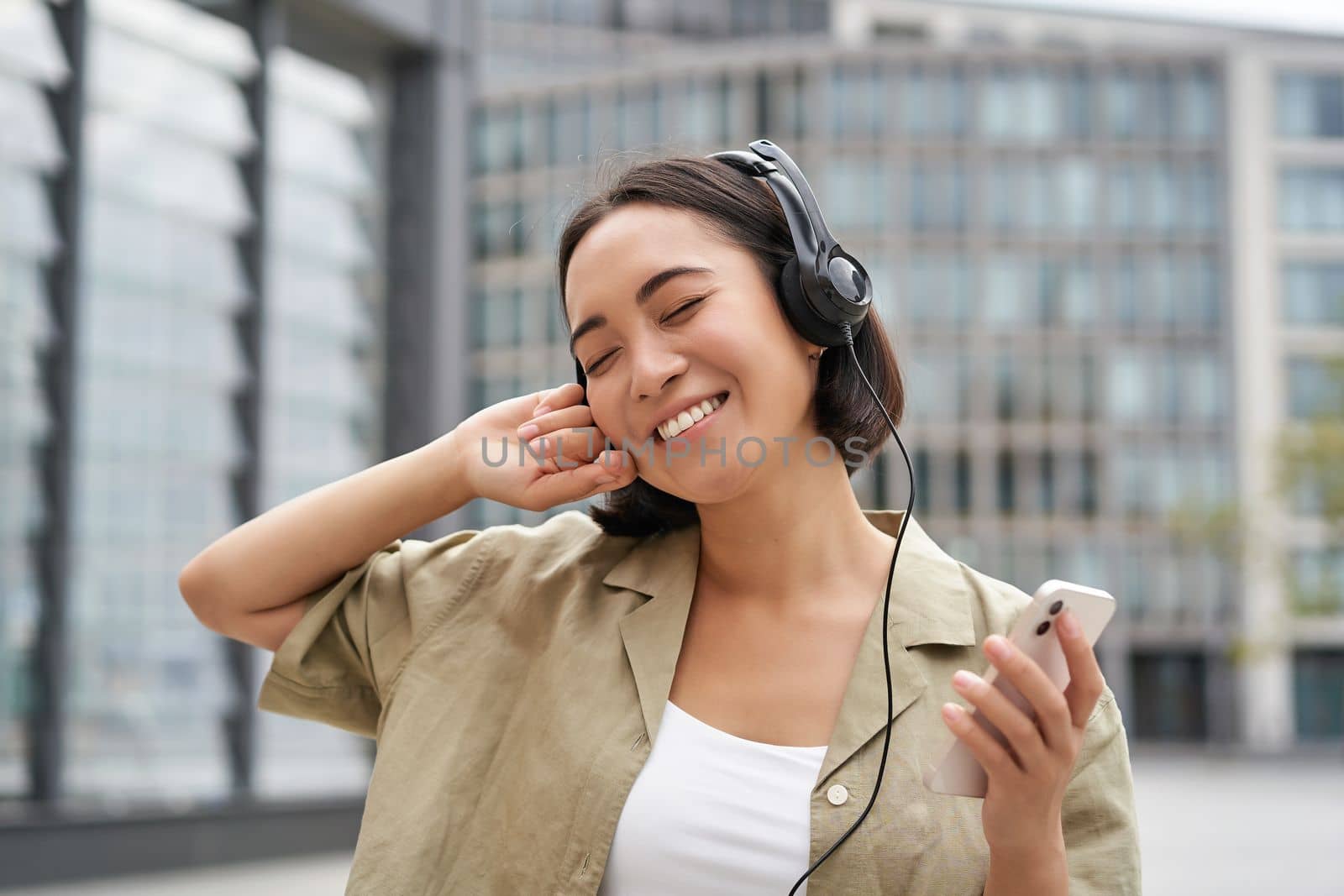 Happy young woman dancing on streets and listening music in headphones, holding smartphone.