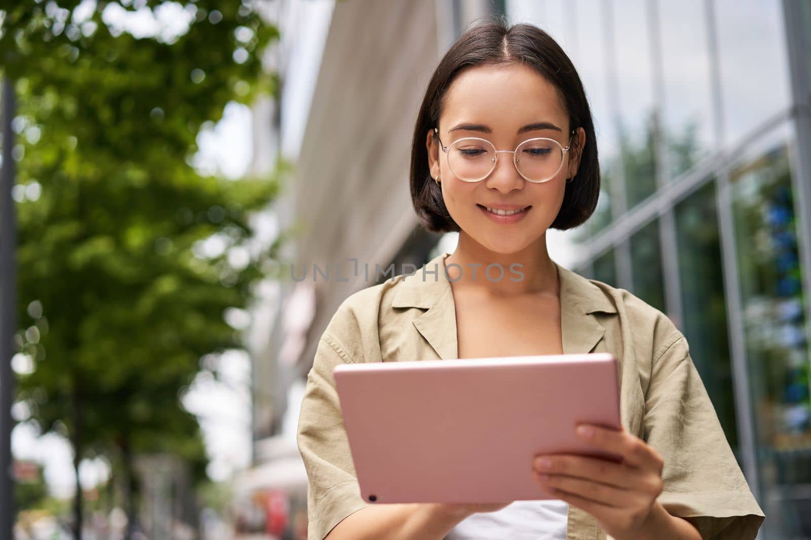 Outdoor shot of young korean woman stands on street with laptop, wears glasses, reads, smiles happily by Benzoix