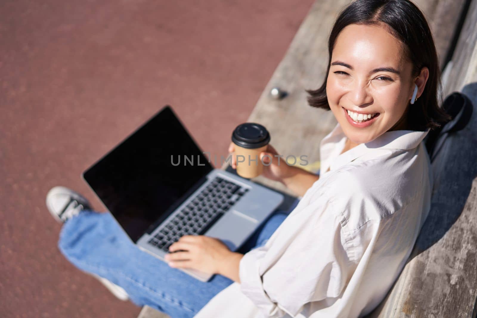 Beautiful young asian female student, sitting with laptop on bench in park, drinking takeaway coffee and listening music, working remotely, studying outdoors.