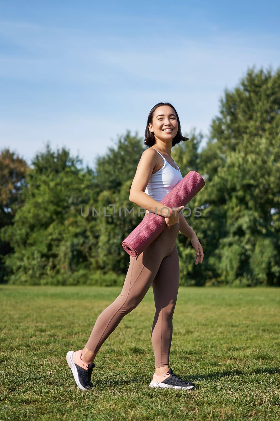 Vertical shot of stylish fitness woman with rubber mat, posing on lawn in park, doing workout on fresh air. Wellbeing and sport concept