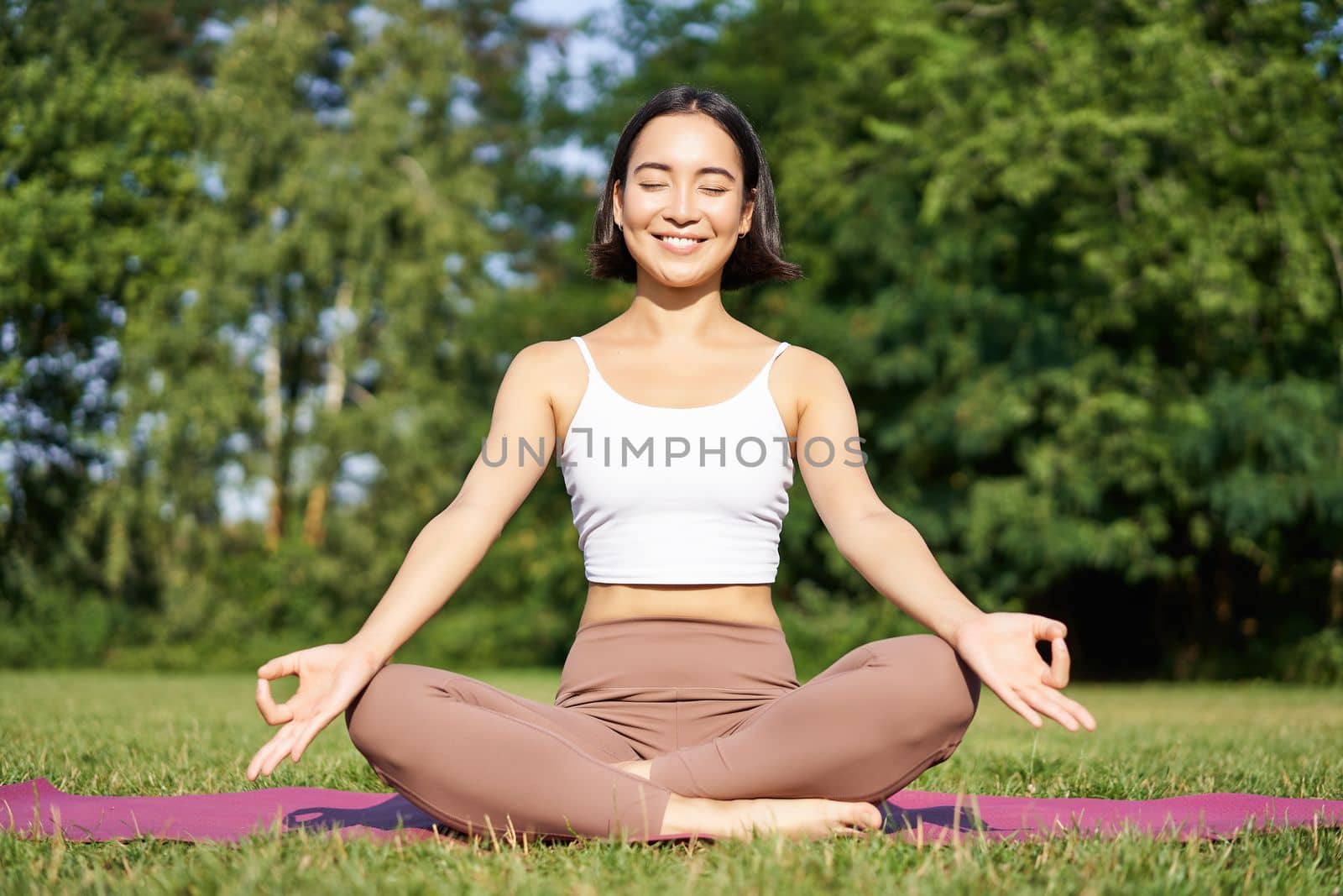 Sporty woman meditating on fresh air, sitting on fitness mat and practice yoga, smiling pleased. Sport and people concept.