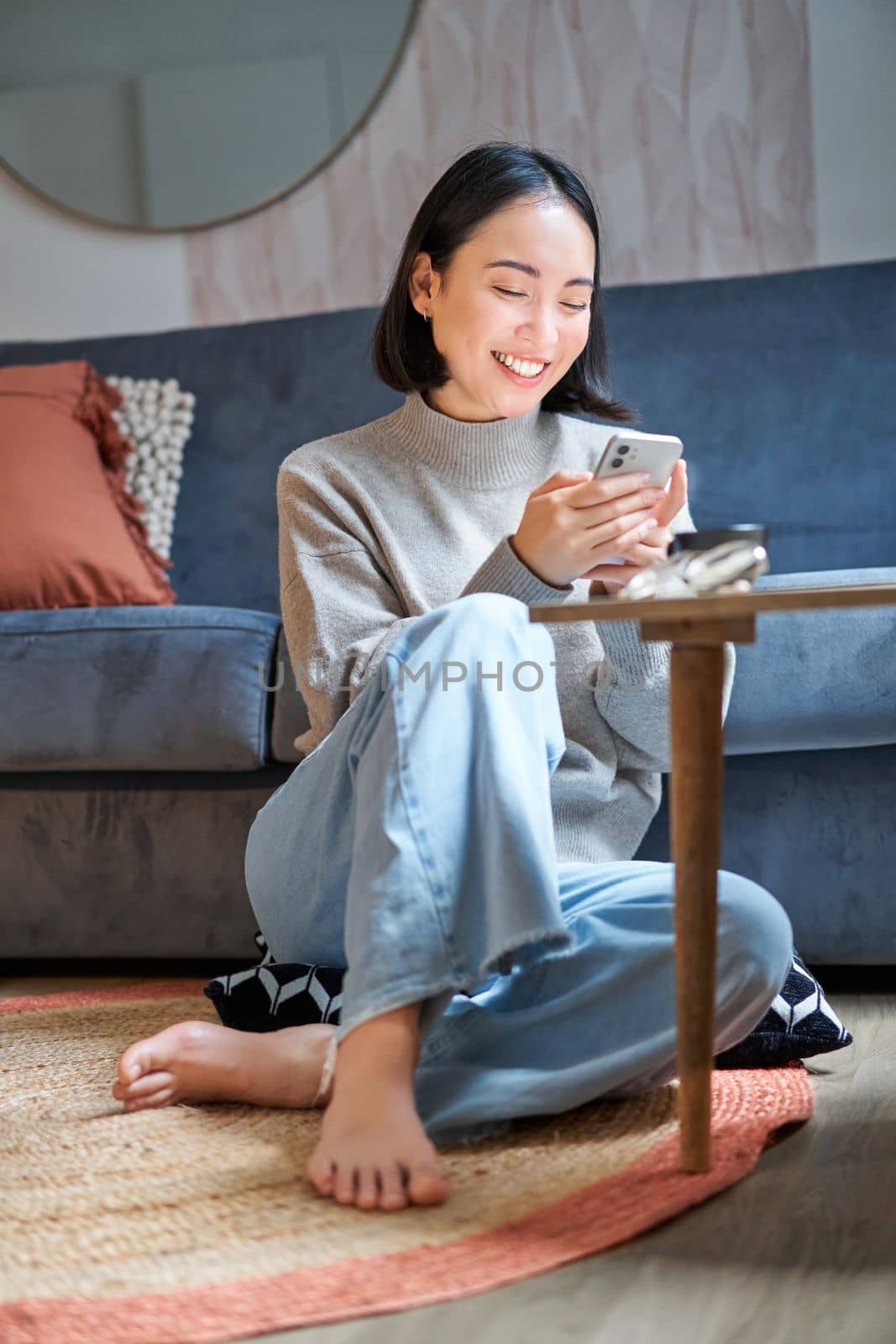 Vertical shot of stylish korean woman sitting on floor at home, using her mobile phone, holding smartphone and smiling.