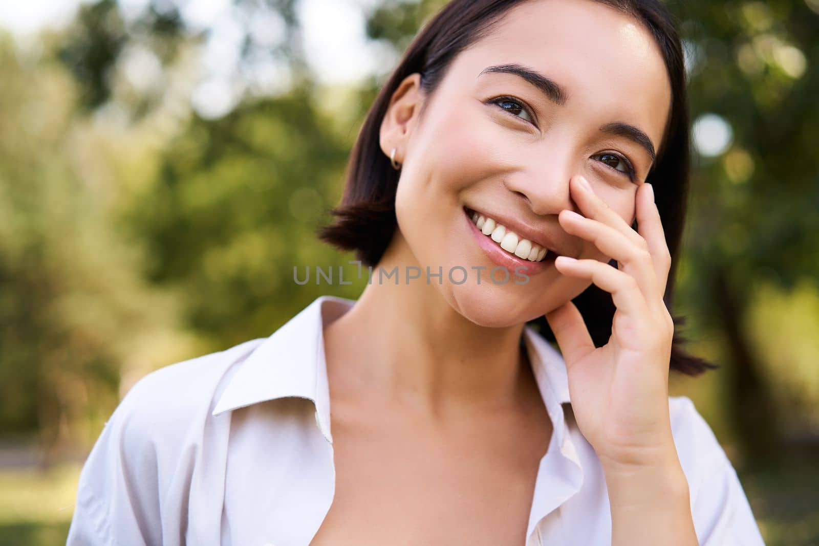 Portrait of young asian woman, laughing and smiling, looking happy while enjoying the walk in park, posing near green trees.