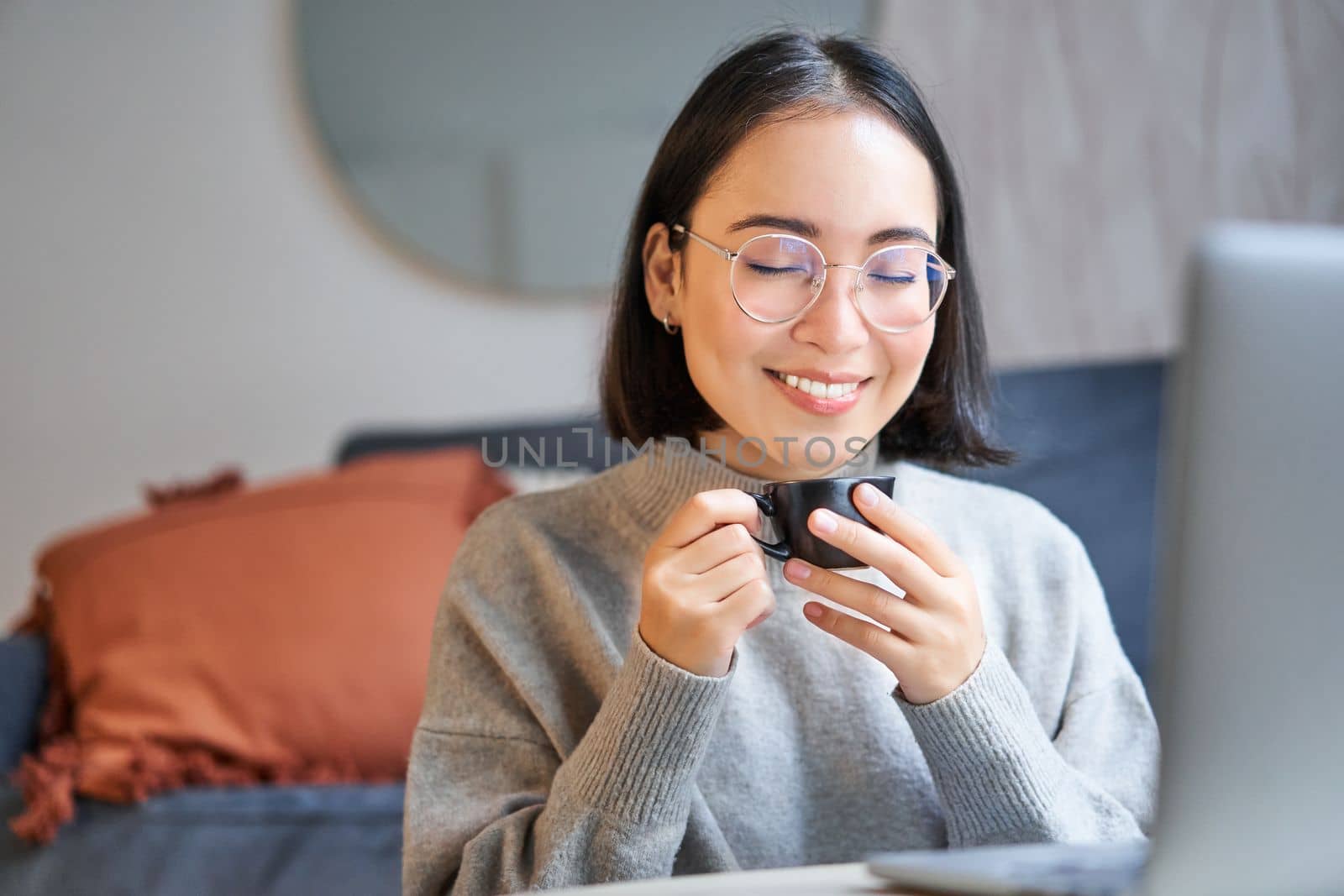 Close up portrait of beautiful asian woman drinks her coffee, smells drink in cup, takes brake from working or studying on laptop.
