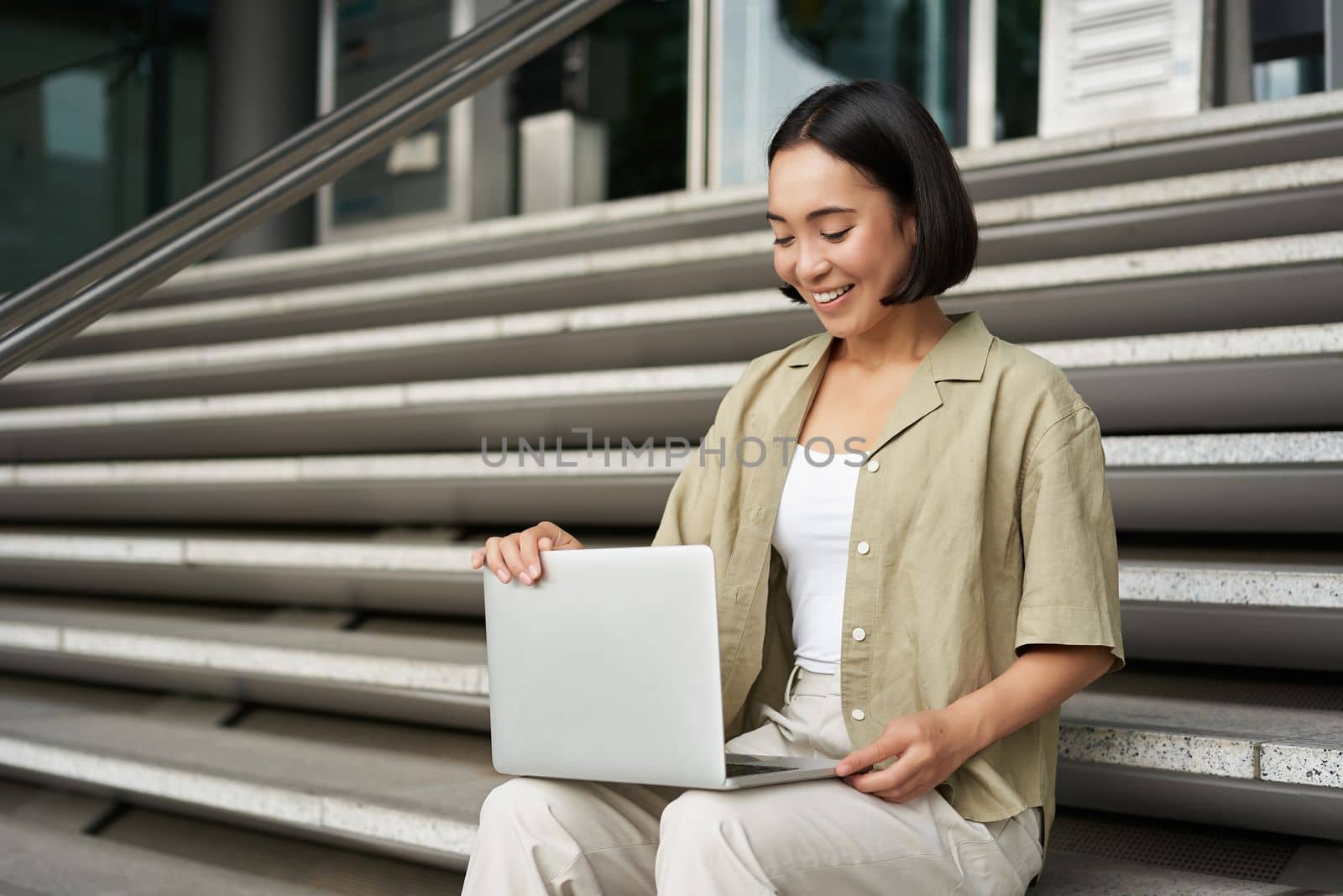 Portrait of young woman student, asian girl using laptop. Asign smiling girl, digital nomad works on her project remotely, sitting on street by Benzoix