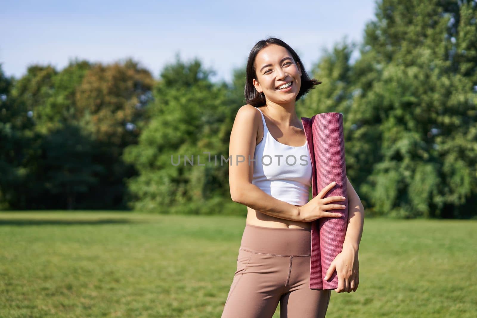 Portrait of asian girl smiles and laughs, stands with sports equipment, rubber mat, wears uniform for workout and exercises in park.